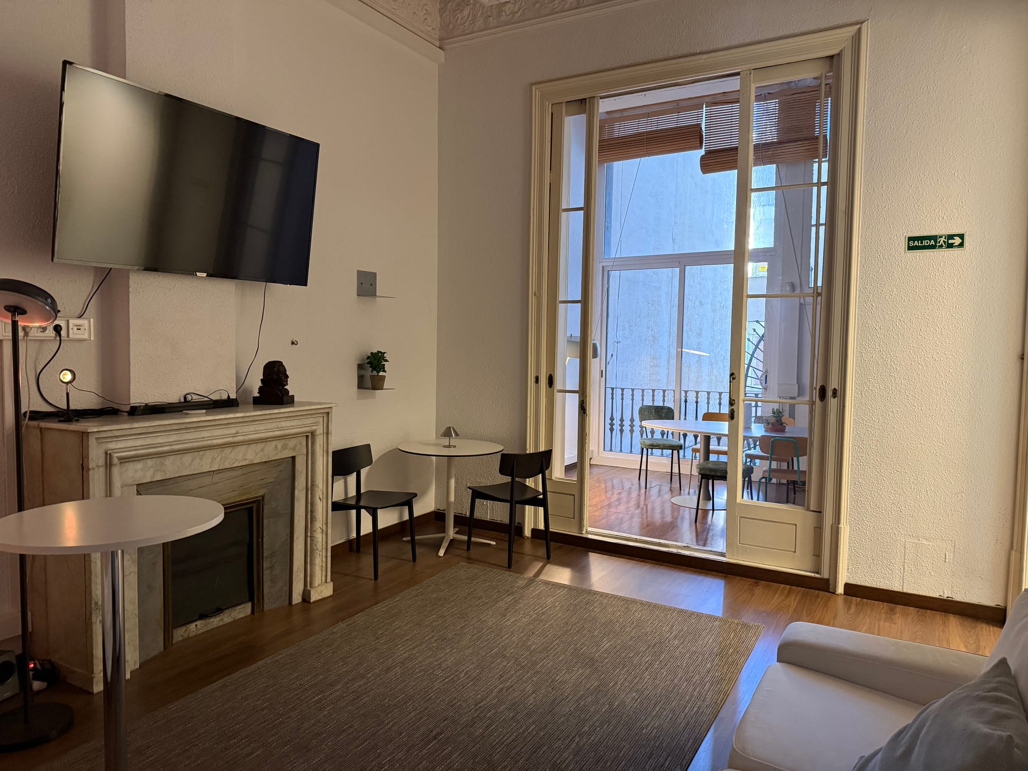 The Eixample 46 lounge: a wall-mounted television, a small fireplace mantel, and chairs by tall French doors opening onto a balcony
