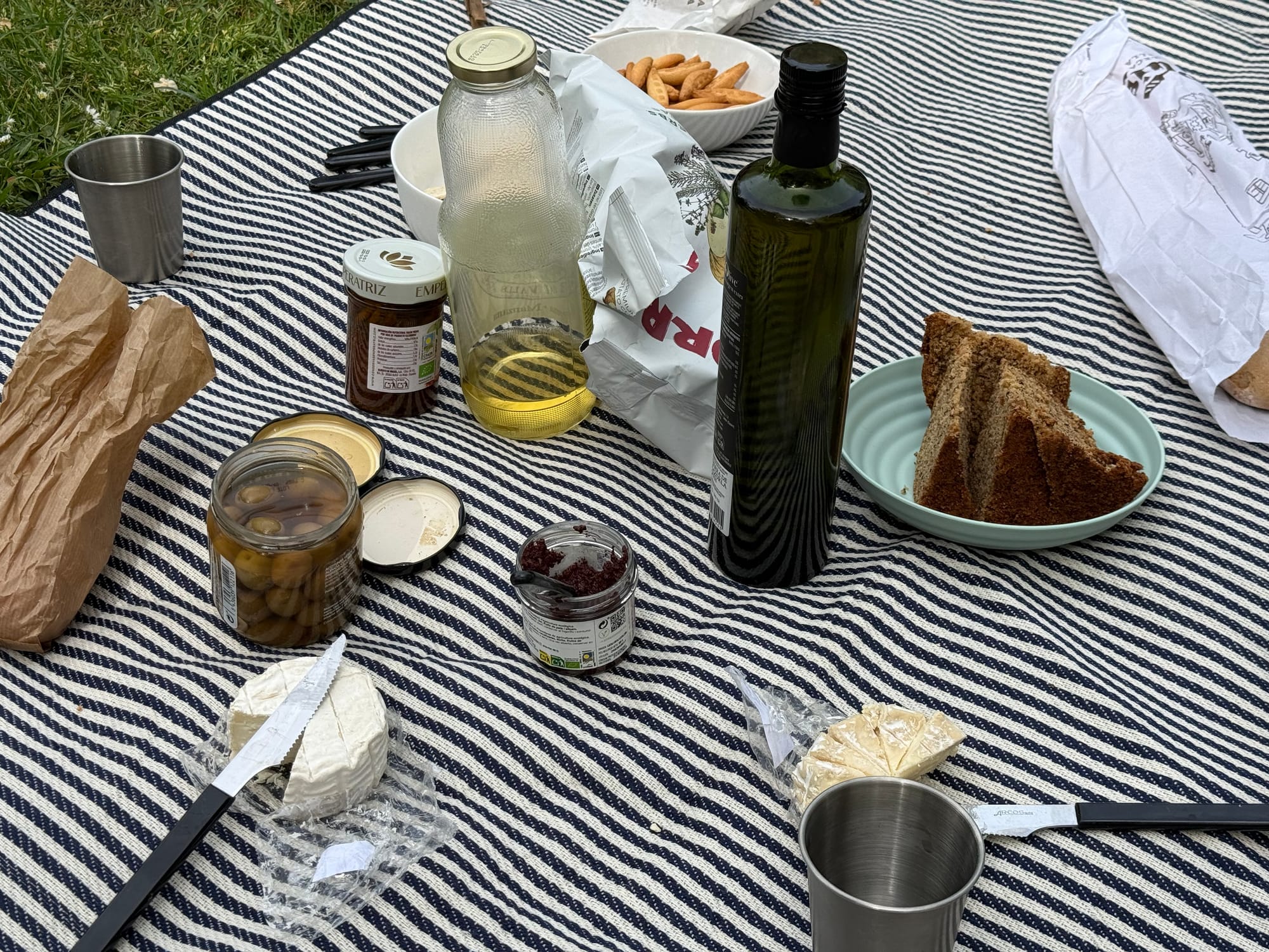A picnic spread on a navy and white striped cloth: bread on a small plate, jars of olives and fig confit, a bottle of olive oil, a glass bottle of apple juice, almonds in a bowl, and small steel cups