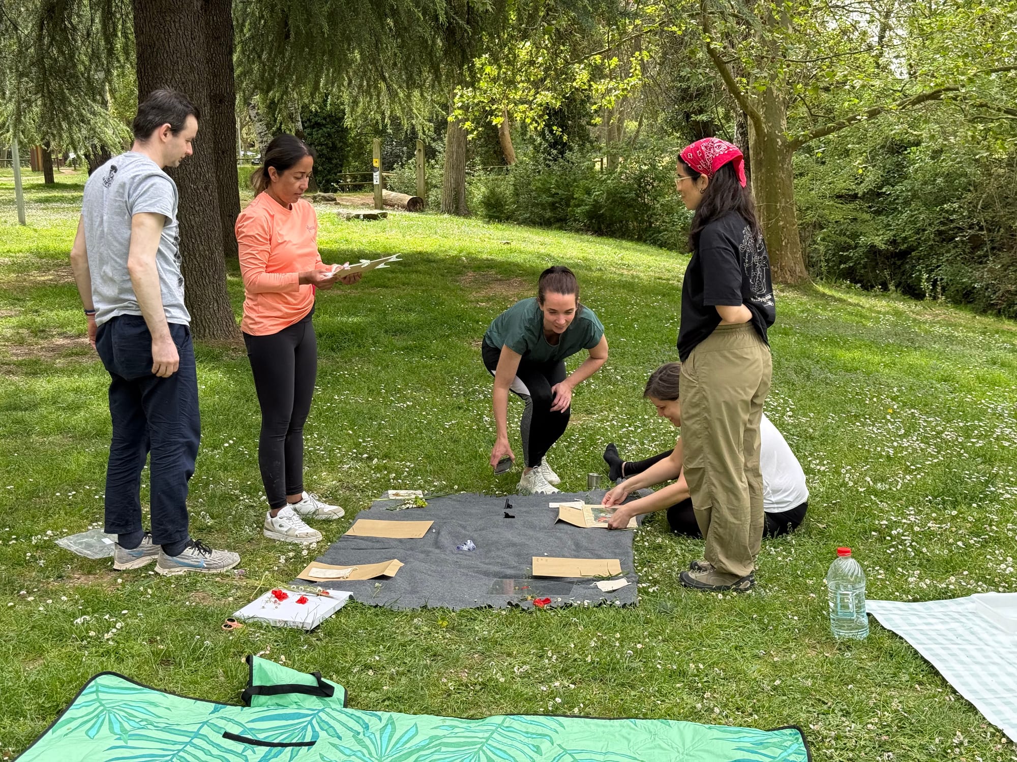 A group of five people standing and crouching around a black blanket on grass in a park, laying out and adjusting cyanotype prints to expose in the sun. Picnic blankets and bags around the edges.