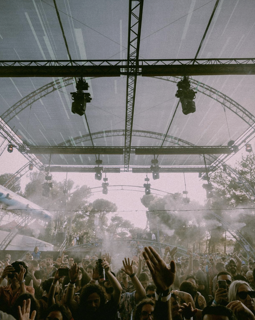 An atmospheric crowd shot inside the Poble Espanyol main tent: a packed audience, hands in the air, dramatic stage lighting and a haze of confetti or smoke