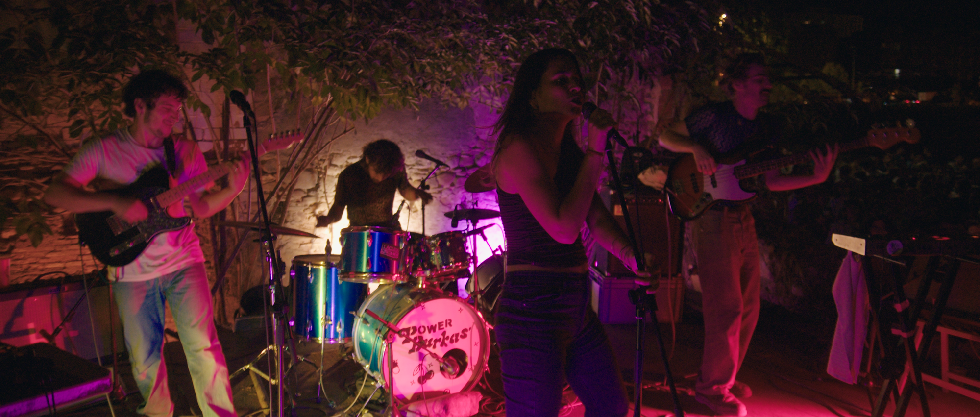 A live rock band performing on an outdoor stage at night under dramatic purple and pink stage lighting, with a vocalist singing into a microphone.
