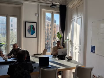 The main Eixample 46 coworking room: a long communal table with members at work, sunlight pouring through tall French windows onto the original wood floor