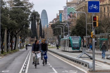 A modern Barcelona tram travels along a street with buildings and trees in the background.