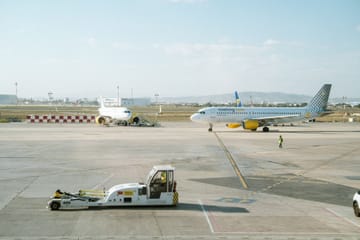 A Vueling aircraft taxiing at Barcelona El Prat Airport under a clear sky, with terminal buildings in the background.