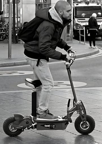 A folded electric scooter standing beside a metro station entrance in Barcelona, with commuters passing by on a weekday morning.