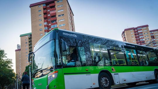 A Moventis bus driving through a street in L'Hospitalet de Llobregat, with buildings and traffic visible in the background.