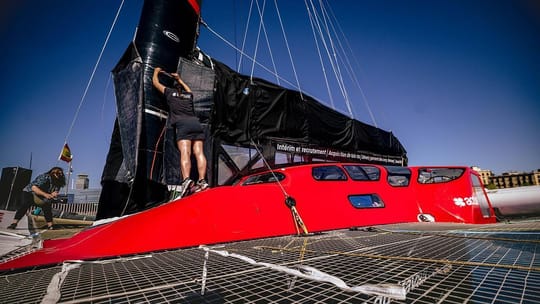 The Actual Ultim 4 trimaran, a large, sleek white boat with three hulls, docked in Barcelona's Marina Port Vell with city bui