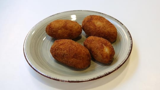 A close-up of golden-brown croquettes on a plate, garnished with fresh herbs, at a food festival.