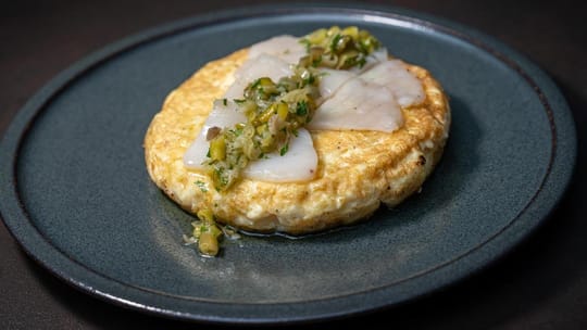 A close-up of a golden, creamy Spanish potato tortilla with a topping, possibly prawn romesco or stewed meat.