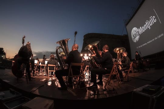 People sitting on a beach at sunset watching a film projected onto a large screen, with the sea in the background.
