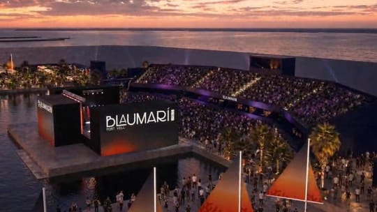 A large floating stage illuminated at night in Barcelona's Port Vell, with city lights and the World Trade Center in the back