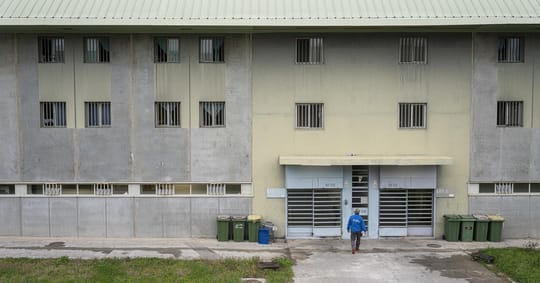 Prison guard standing in a corridor of a modern prison facility, looking towards the camera with a serious expression.