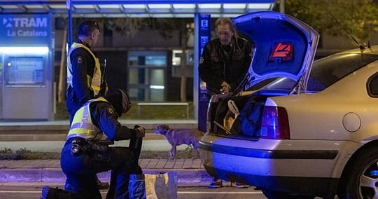 Mossos d'Esquadra officers on patrol in a city street, with a police vehicle in the background.
