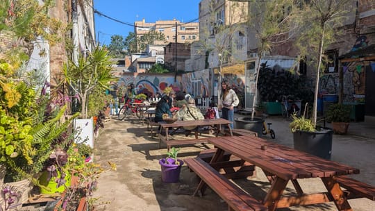 A vibrant community garden in Poblenou, Barcelona, with lush greenery, wooden tables, and residents enjoying the space.