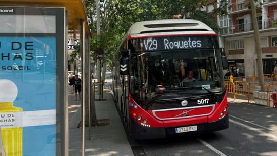 A modern articulated TMB bus drives along a sunny street in Barcelona with people walking nearby.