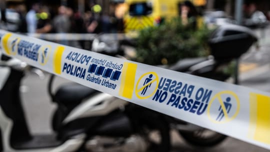 Police tape blocks a street in Barcelona, with emergency vehicles in the background, indicating a crime scene.