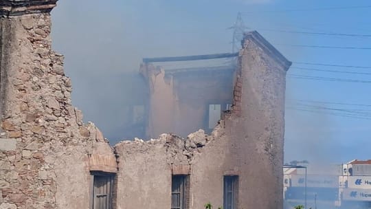 Smoke rising from the roof of Torre Gran, a large, old stone farmhouse with a damaged roof, surrounded by vegetation.