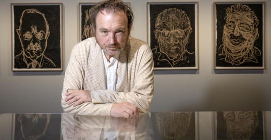 Miquel Barceló, an artist with dishevelled hair, stands in front of his artwork at an exhibition.