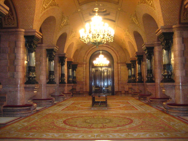 A long, opulent hall with a high arched ceiling, multiple chandeliers, and a richly patterned carpet. Ornate columns line the walls.