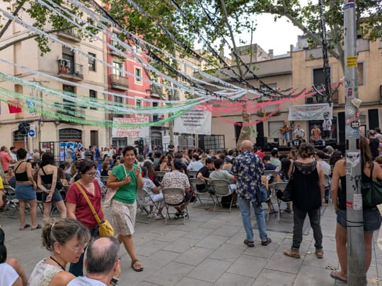 Crowd at a decorated street in Gràcia during the 2025 festival, with a bar set up on the pavement serving drinks under fairy lights.