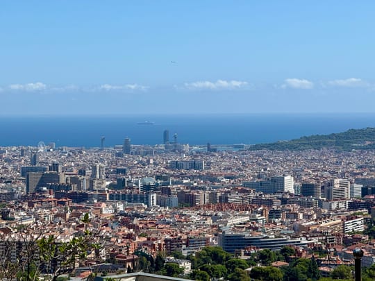 A panoramic view of the Collserola Natural Park bordering Barcelona, with dry summer vegetation and warning signs about forest fire risks visible along a trail.