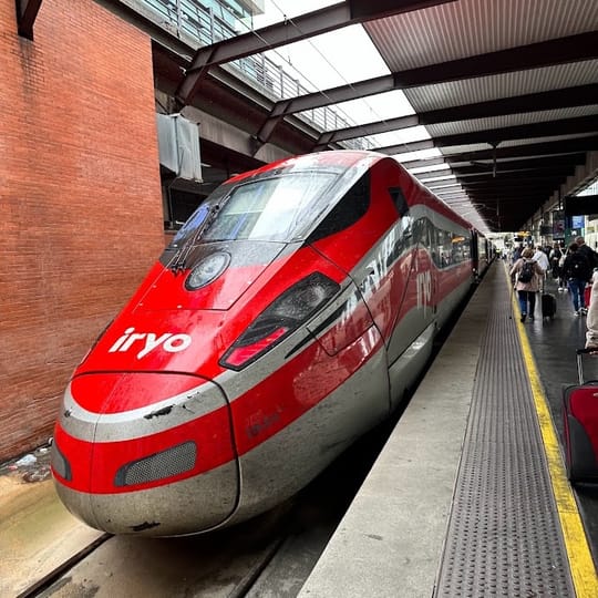A red and grey Iryo high-speed train is parked at a station platform. Passengers with luggage are visible further down the platform.