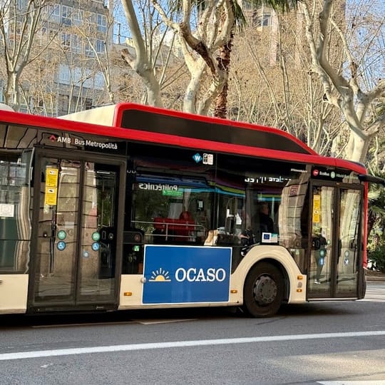 A red and cream public bus with an 'OCASO' advertisement on its side drives along a city street lined with trees and buildings.