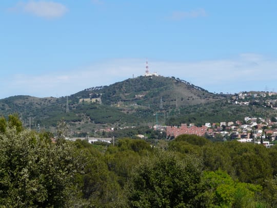 Smoke rising from the Collserola mountains near Barcelona, with emergency vehicles and a Guardia Urbana car blocking access to a forest road.