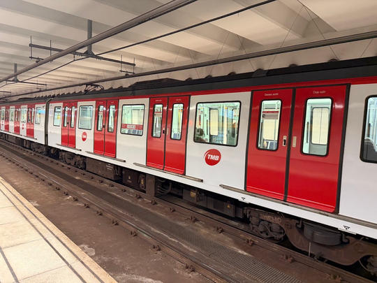 A red and white metro train with 'TMB' logo on its side is stopped at an underground station platform. The train's doors are closed, and the tracks are visible in the foreground.