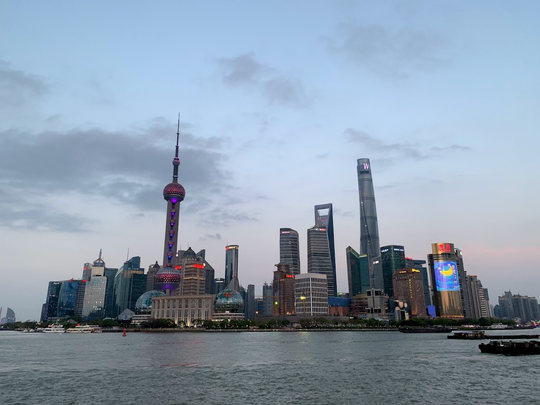 A wide shot of the Shanghai skyline at dusk, with the Oriental Pearl TV Tower prominent among numerous illuminated skyscrapers lining a river.