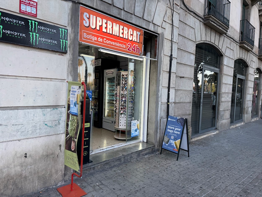 A 24-hour convenience store entrance with an orange 'Supermercat' sign. Monster Energy logos are visible on the left wall, and a 'Store your bags here' sign is on the pavement.