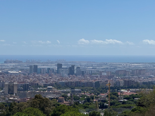 A wide shot of a city with many buildings and a port in the distance, leading to the sea under a blue sky with scattered clouds.