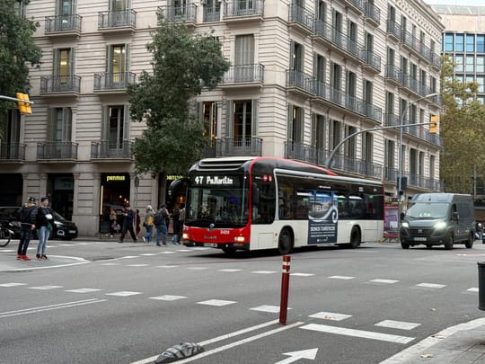 A bus stops at the UAB campus in Bellaterra, surrounded by trees and university buildings, with students boarding under a sheltered stop.