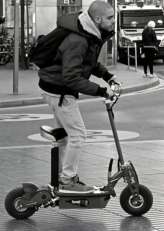 A folded electric scooter standing beside a metro station entrance in Barcelona, with commuters passing by on a weekday morning.