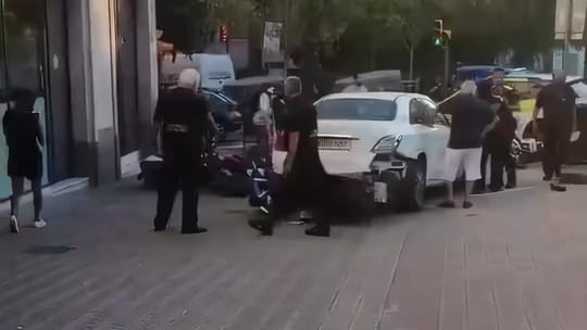 A white Mercedes stopped on the pavement beside damaged motorcycles on carrer de Jordi Girona, Pedralbes, with Guàrdia Urbana officers at the scene
