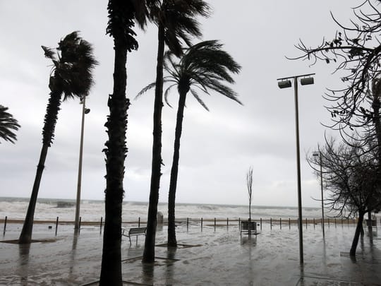 On the seafront promenade in Barceloneta, palm trees are bent by strong winds, and the grey sky hangs over rough, white-capped waves.