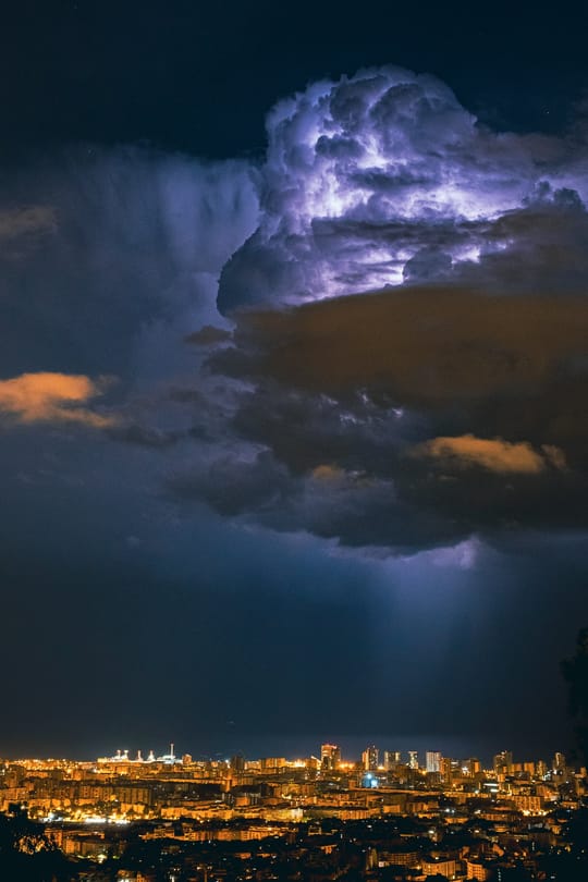 A huge thunderstorm cloud, lit from within by lightning, towers over the illuminated skyline of Barcelona at night.