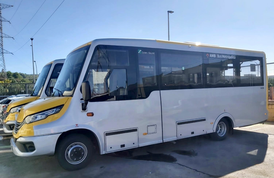 A modern AMB minibus on a quiet residential street in Molins de Rei, surrounded by green hills and low-rise housing.