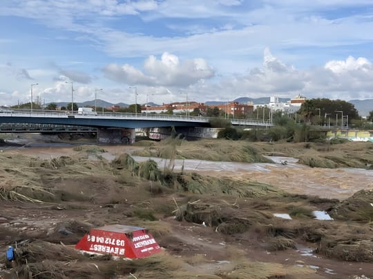 Aerial view of the Besòs river park showing flood debris piled near the Rodalies railway bridge, with partially cleared walkways on the left bank.