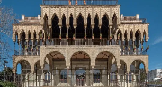 A historic Neo-Mudéjar building, Casa de les Altures, with intricate brickwork and archways, surrounded by green trees under 
