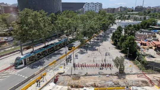 A modern tram travelling along a street in Barcelona, with buildings and trees in the background under a clear sky.