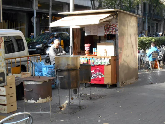 A street vendor roasts chestnuts in a brazier on a Barcelona pavement during autumn, with fallen leaves and city buildings in the background.