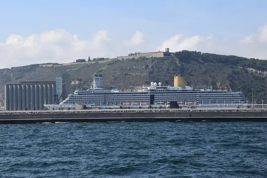A cruise ship docked at the Port of Barcelona near Pont d'Europa, with city skyline in the background.