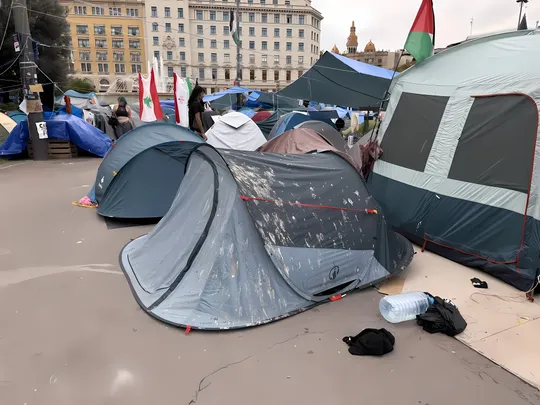Aerial view of a large encampment with hundreds of tents in Plaça Catalunya, Barcelona, surrounded by city buildings and protest banners.