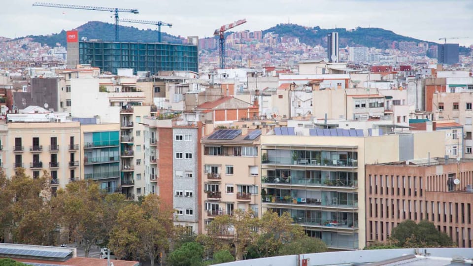 A 'For Rent' sign displayed in a window in Barcelona, with a blurred street scene in the background, symbolising the city's r