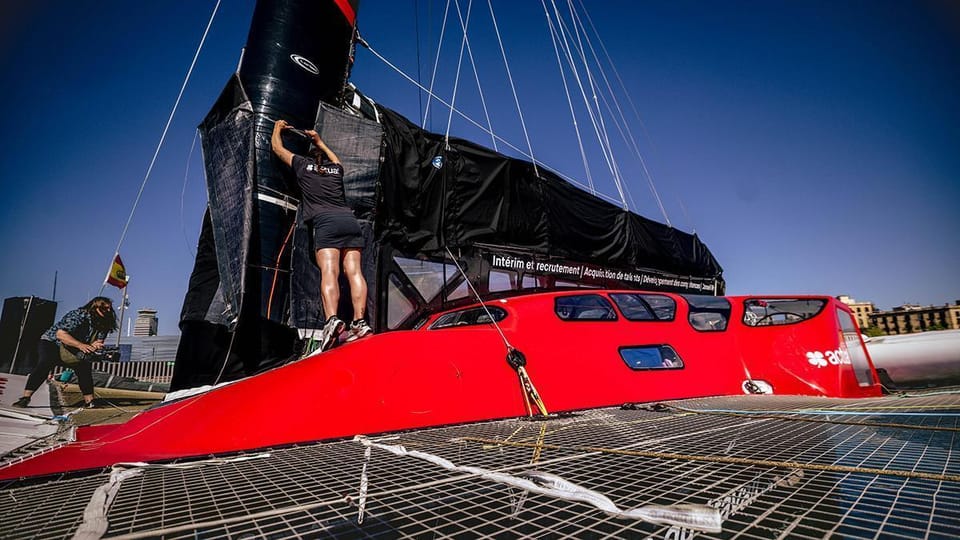 The Actual Ultim 4 trimaran, a large, sleek white boat with three hulls, docked in Barcelona's Marina Port Vell with city bui