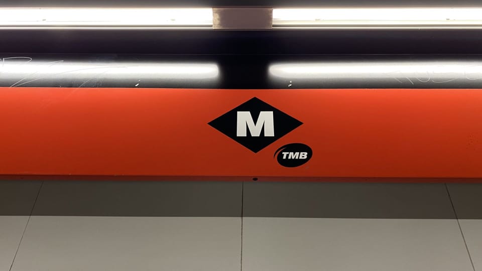 A Barcelona Metro train arriving at a station platform, with a digital display showing service information.