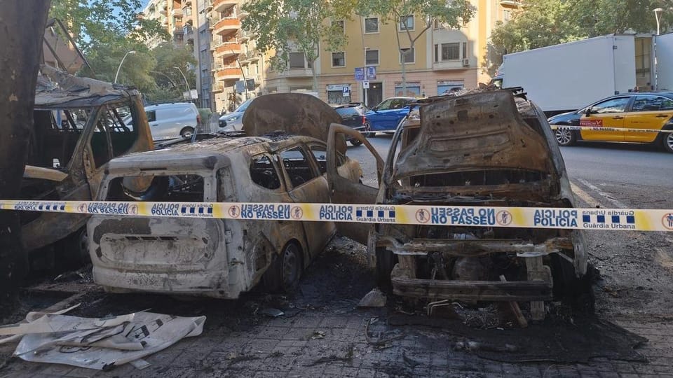 Burnt vehicles and a damaged traffic light control cabinet on a street corner in Barcelona's Eixample district.