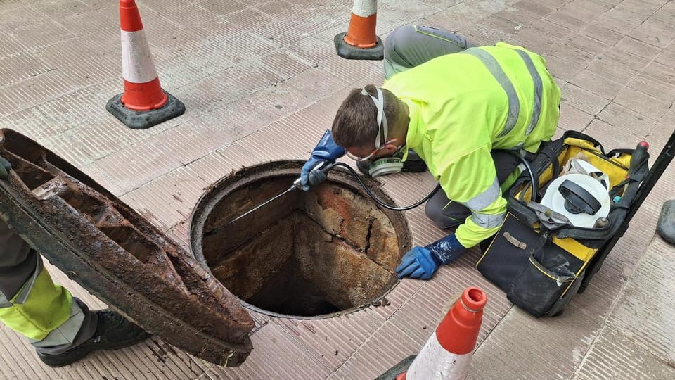 A worker in protective gear inspects a sewage manhole in an urban street, possibly applying pest control treatments.