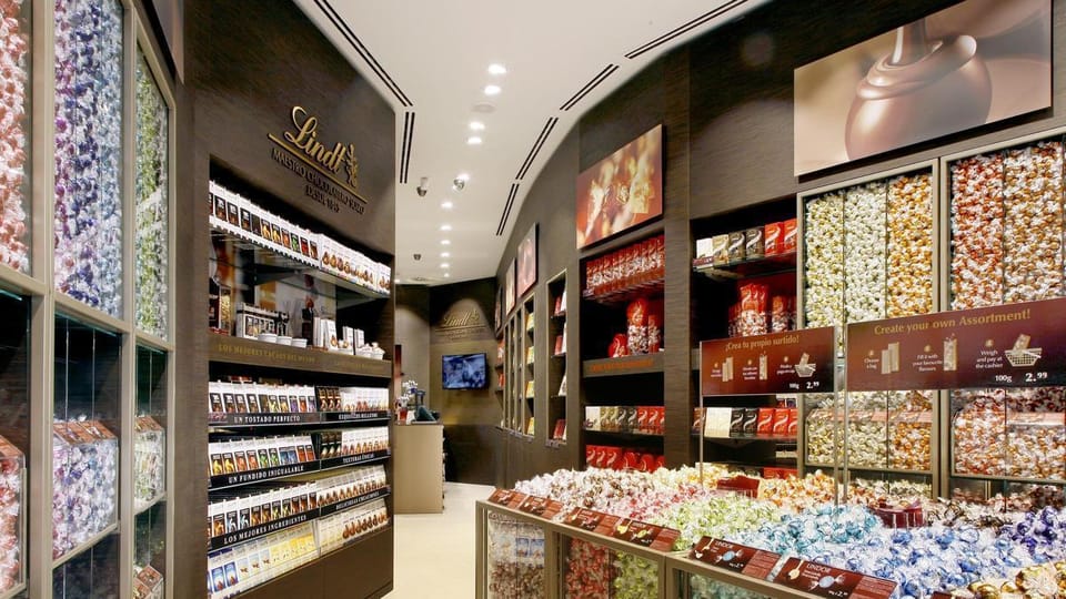 Exterior view of a Lindt chocolate shop on a tree-lined street in Barcelona, with people walking past.